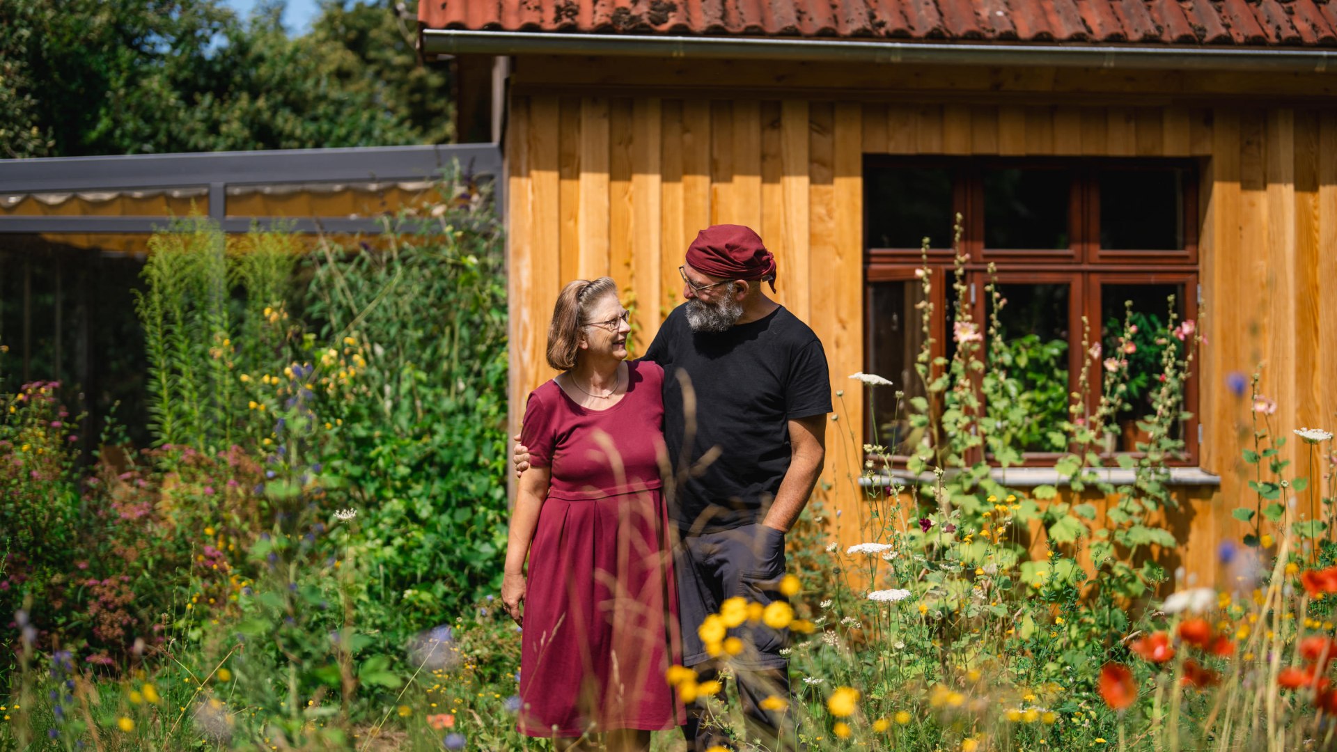 A couple is standing in a blooming garden in front of a wooden house, smiling at each other, with colorful wildflowers in the foreground.