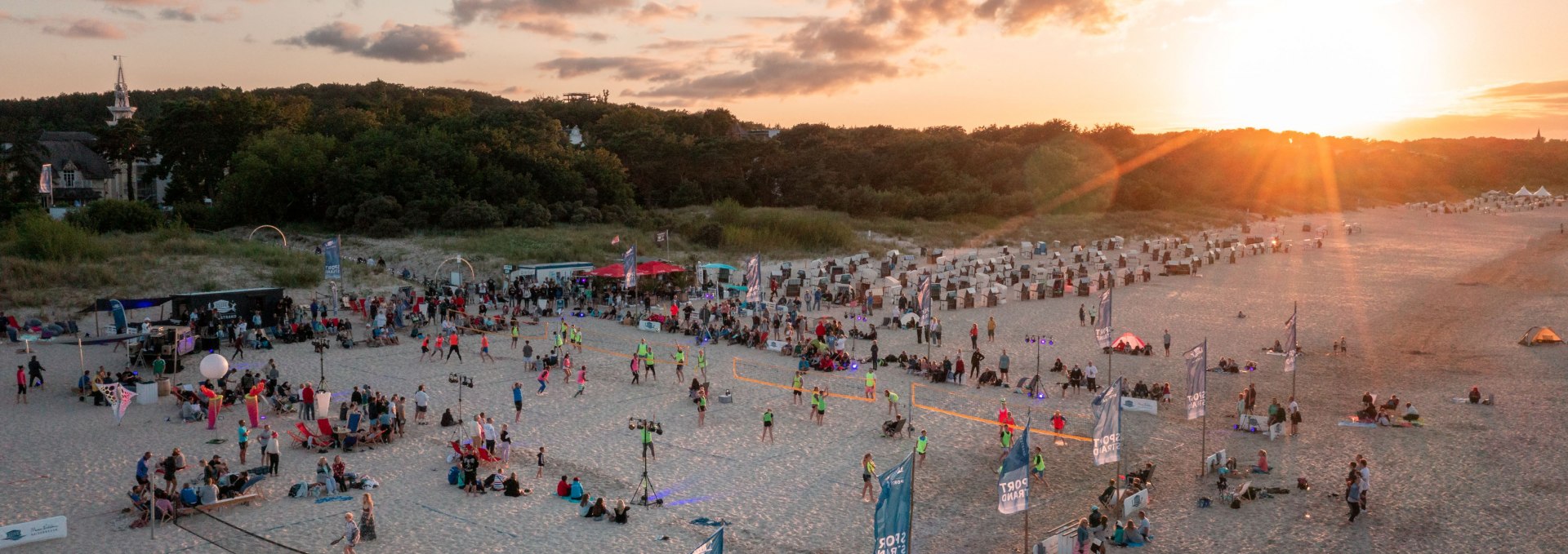 Mensen spelen glowvolleybal bij zonsondergang op het Oostzeestrand van de Kaiserbäder tegen een achtergrond van duinen en strandstoelen.