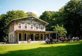 Ein helles Geb&auml;ude mit einer Terrasse, vor dem mehrere Personen auf B&auml;nken auf einer gr&uuml;nen Wiese sitzen. // &copy; Nationalpark-Zentrum K&Ouml;NIGSSTUHL / Dan Petermann