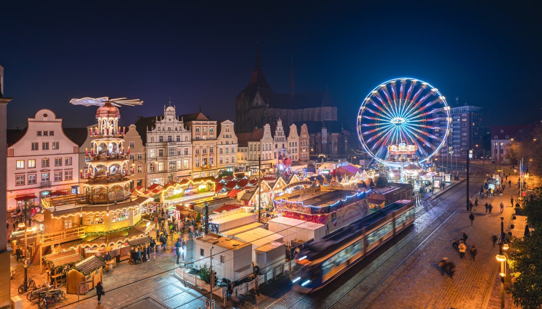 Feestelijke lichtjes in de Hanzestad - de kerstmarkt van Rostock betovert met een reuzenrad, historische ambiance en culinaire hoogstandjes op de Neuer Markt., © TMV/Gross Uitzicht op de feestelijk verlichte kerstmarkt in Rostock met reuzenrad en historische gebouwen bij nacht.