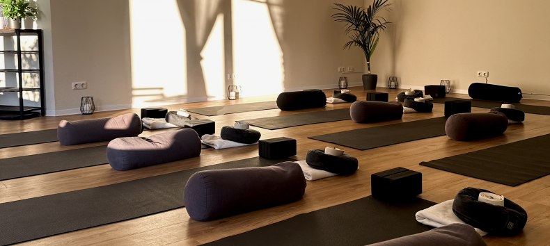 Bright, minimalist yoga room with wooden flooring, several yoga mats, meditation cushions, bolsters and yoga blocks. Warm sunlight shines through large windows and creates a calm, relaxing atmosphere for yoga, meditation and relaxation. // &copy; MOVA PLACE - Yoga & Co. Studio