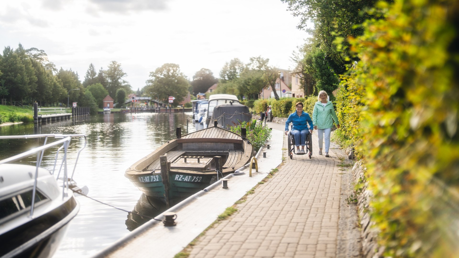 A wheelchair user out and about with her friend on the canal in Plau am See. Boats are moored at the edge.