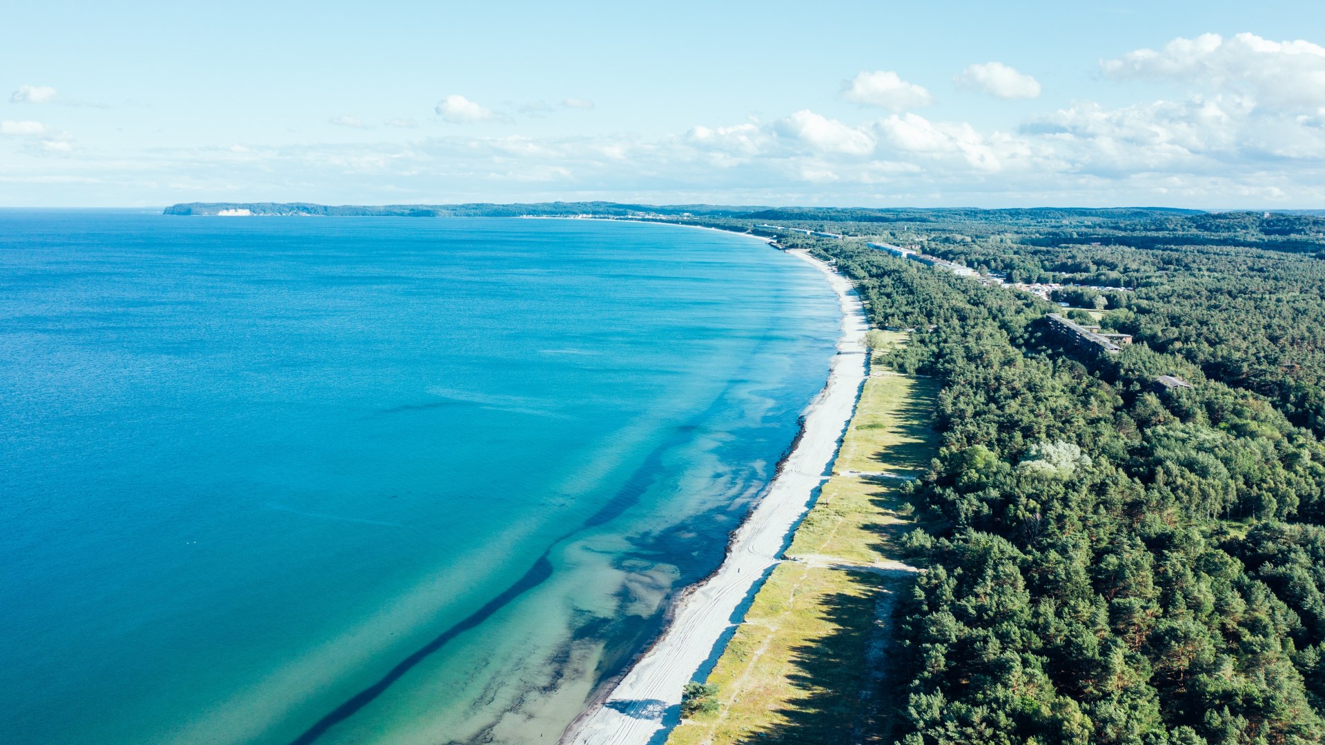 Aerial view of the Baltic Sea coast on R&uuml;gen with clear turquoise blue water, light-colored sandy beach and adjacent coastal forest.