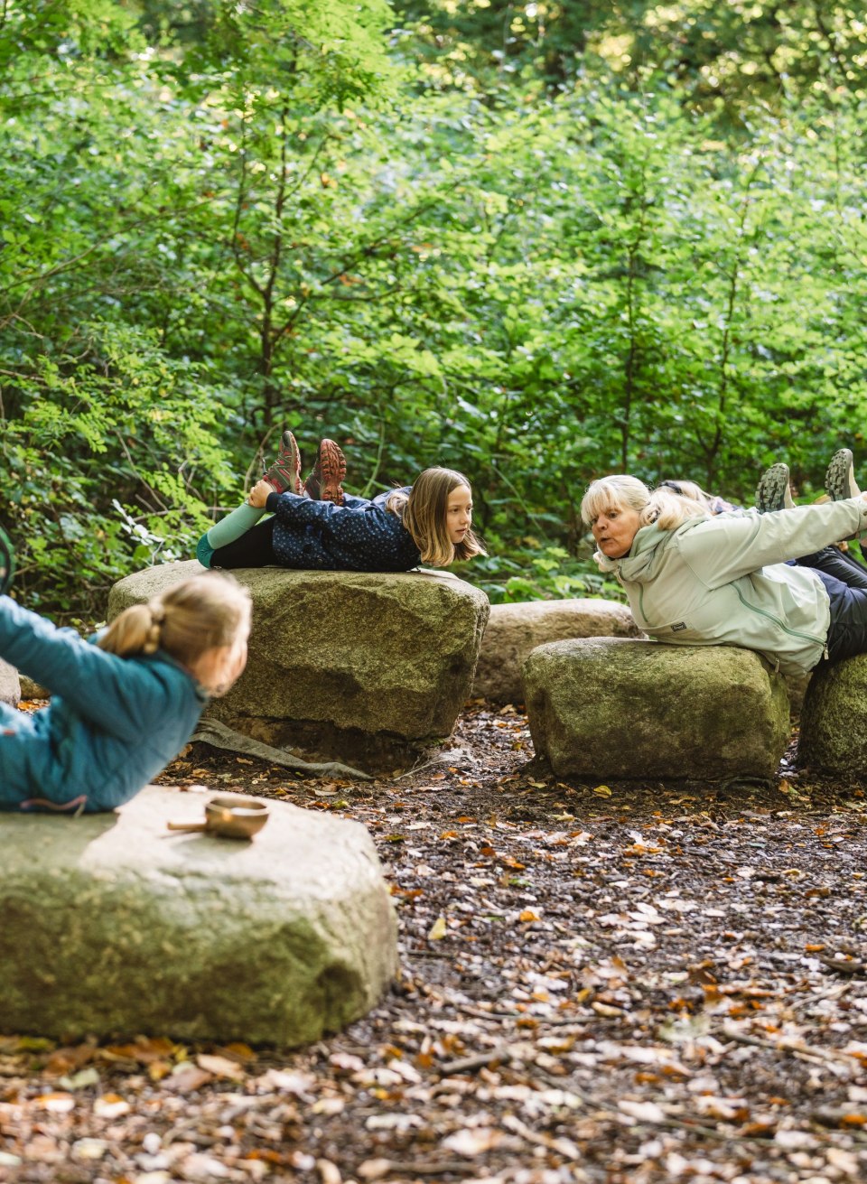 Kinderen en een volwassene liggen op grote stenen in het bos en doen samen yoga-oefeningen, omringd door dichte bomen en bladeren.