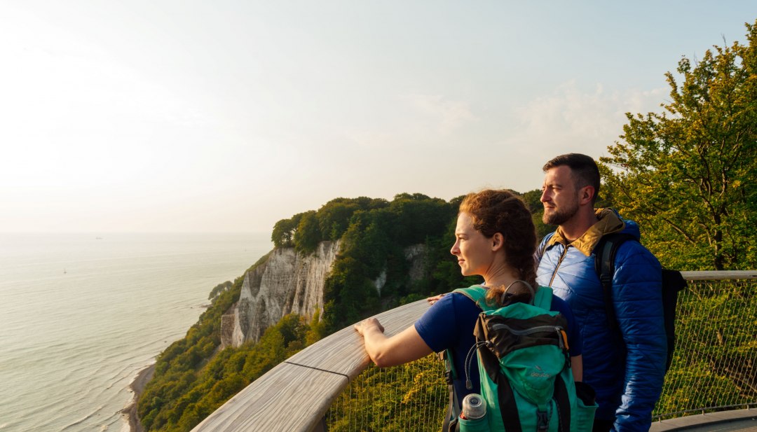 Magiczny widok na kredowe wybrzeże Rugii - para wędrowców podziwia zachód słońca nad Morzem Bałtyckim z punktu widokowego w Parku Narodowym Jasmund., © TMV/Petermann Dwóch wędrowców spogląda na kredowe klify i Morze Bałtyckie w Parku Narodowym Jasmund na wyspie Rugia.