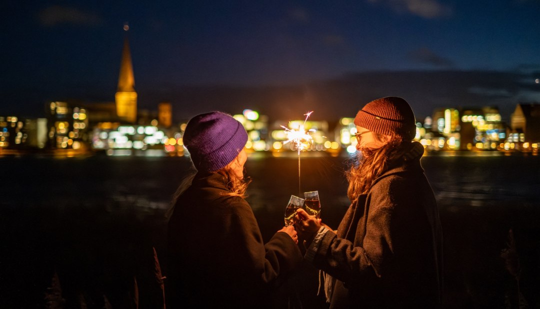 Feestelijke oudejaarsavond - fonkelende lichtjes tegen de achtergrond van Rostock in Mecklenburg-Vorpommern., © TMV/Witzel Twee mensen proosten met mousserende wijn terwijl ze een sparkler vasthouden, met de verlichte skyline van Rostock in Mecklenburg-Vorpommern op de achtergrond.
