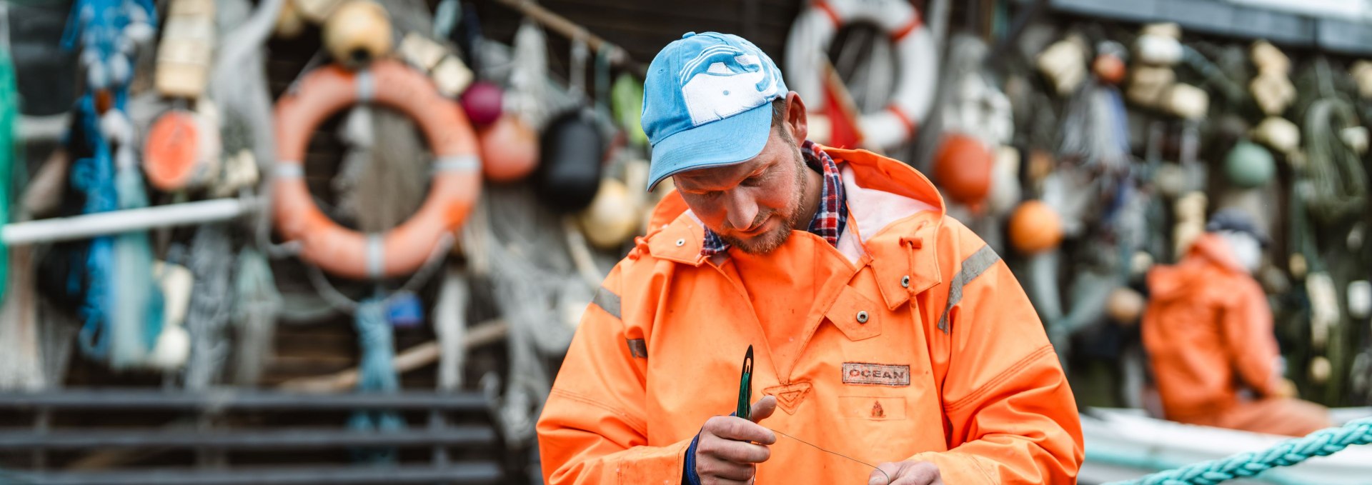 Fisherman in the harbor of Gager on the island of R&uuml;gen repairs a net in front of a hut with lifebuoys and fishing gear, while a colleague sits in the background.