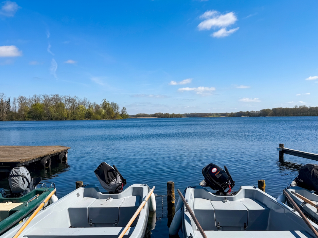Uitzicht vanaf de aanlegsteiger over het Krakower Meer bij stralend zonlicht. Op de voorgrond liggen verschillende roeiboten en motorboten van de bootverhuur van het vakantiepark Kiefernhain aan de steiger; op de achtergrond strekt zich het uitgestrekte, rustige wateroppervlak uit met beboste oevers. // © Feriensiedlung Kiefernhain Uitzicht vanaf de aanlegsteiger over het Krakower Meer bij stralend zonlicht. Op de voorgrond liggen verschillende roeiboten en motorboten van de bootverhuur van het vakantiepark Kiefernhain aan de steiger; op de achtergrond strekt zich het uitgestrekte, rustige wateroppervlak uit met beboste oevers. // © Feriensiedlung Kiefernhain