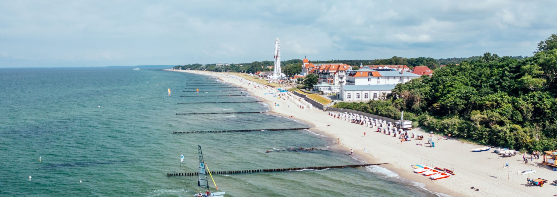 Widok z lotu ptaka na plażę w K&uuml;hlungsborn z ostrogami, leżakami, diabelskim młynem i żagl&oacute;wką nad Morzem Bałtyckim.