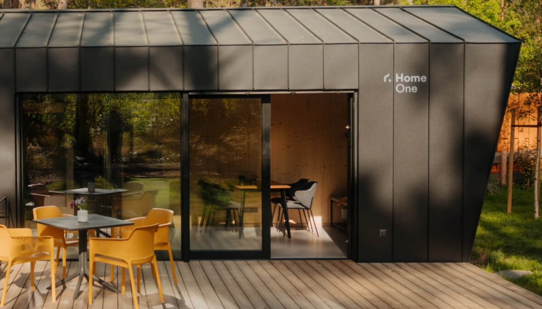 Modern tiny house met donkere gevel, groot houten terras en buitenzitje, idyllisch gelegen in het bos.