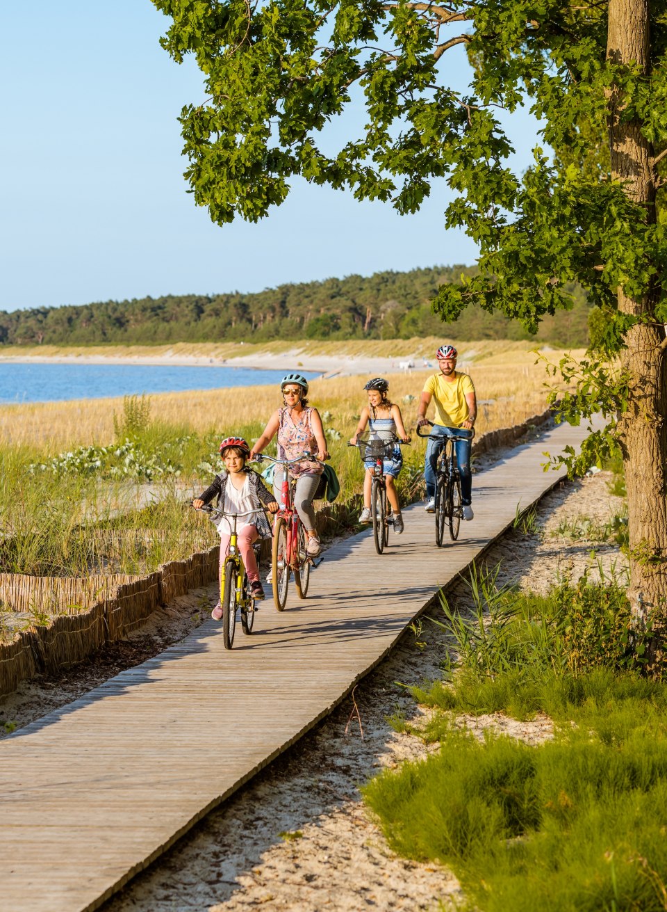 Een gezin fietst over een houten pad op het strand van de badplaats Lubmin, omgeven door duinen, gras en uitzicht op het kalme water van de Bodden.