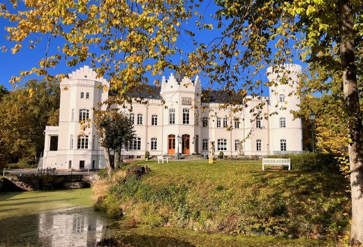 Voor kasteel Schlemmin schitteren herfstbladeren boven de gracht; de witte gevel steekt af tegen de blauwe lucht. // De witte gevel van het kasteel, het goudkleurige gebladerte, de stille slotgracht – Kasteel Schlemmin laat Mecklenburg-Vorpommern van zijn heldere kant zien. // © Schloss Schlemmin Voor kasteel Schlemmin schitteren herfstbladeren boven de gracht; de witte gevel steekt af tegen de blauwe lucht.