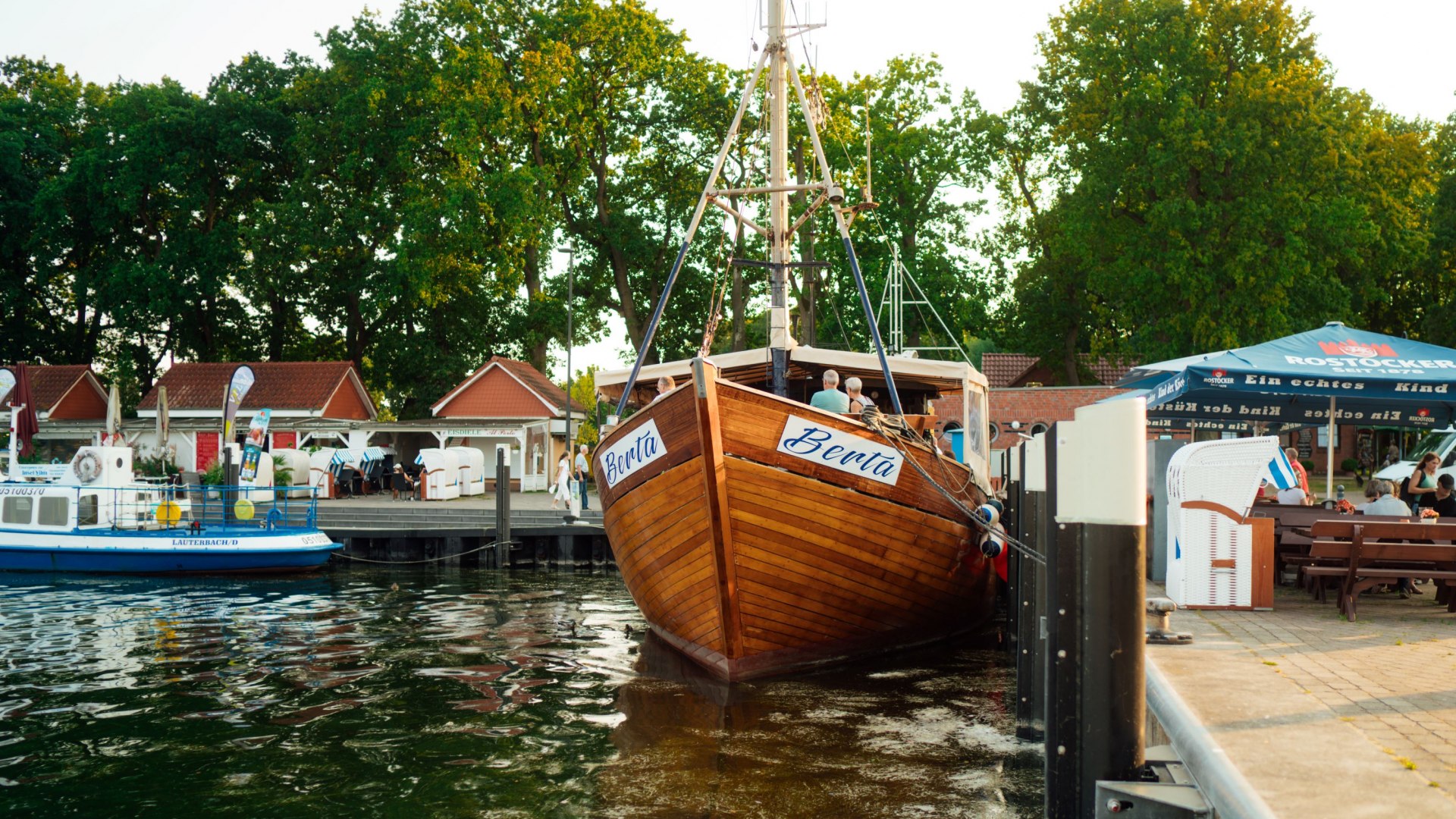 Een houten kotter met de naam "Berta" ligt afgemeerd in de haven van Lauterbach op het eiland Rügen. Kleine rode gebouwen, strandstoelen en zitjes onder parasols zijn zichtbaar op de achtergrond. Omgeven door rustig water en groene bomen biedt het landschap een idyllische havensfeer.
