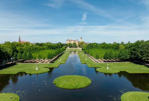 Schwerin baroque garden with symmetrical canal, water features and castle in the background under a blue sky. // Schwerin's palace garden invites you to take a deep breath - baroque water features reflect the fairytale palace, while avenues of lime trees provide shade. Discover where architecture and nature merge harmoniously. // © MV-T/Gänsicke Schwerin baroque garden with symmetrical canal, water features and castle in the background under a blue sky.