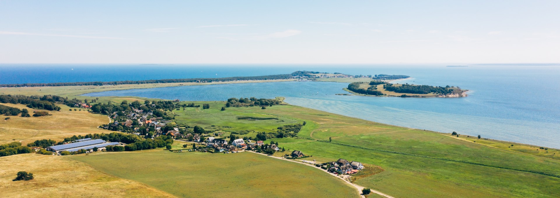 Aerial view of the Zicker mountains on the M&ouml;nchgut peninsula with wide meadows, small villages and a view of the Baltic Sea and surrounding peninsulas under a clear sky.