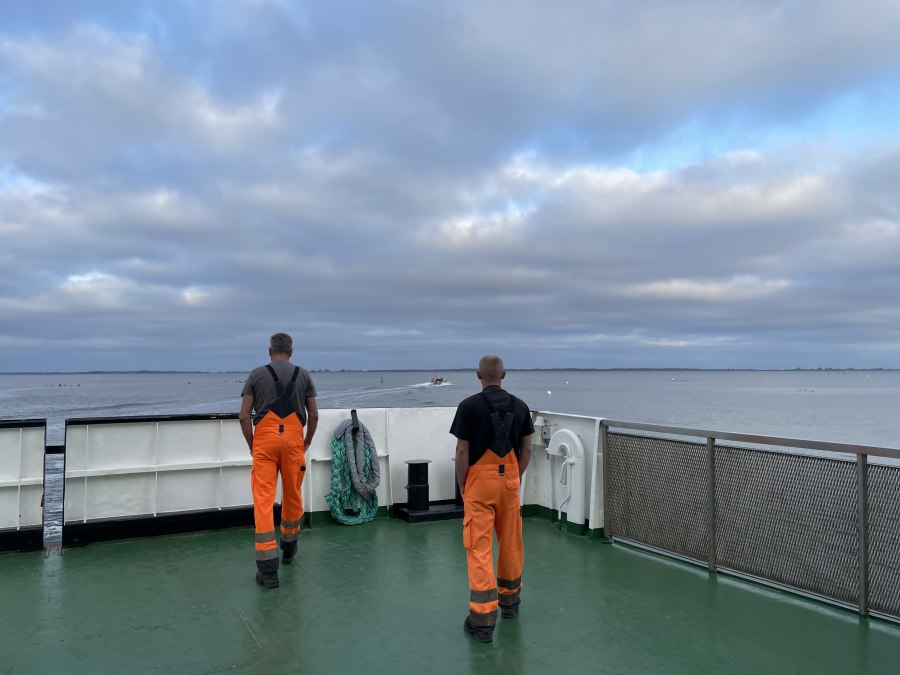 Na promie dwóch pracowników w pomarańczowych ogrodniczkach stoi przy barierce i spogląda na wodę, © Anja Heitmüller Na promie dwóch pracowników w pomarańczowych ogrodniczkach stoi przy barierce i spogląda na wodę, © Anja Heitmüller
