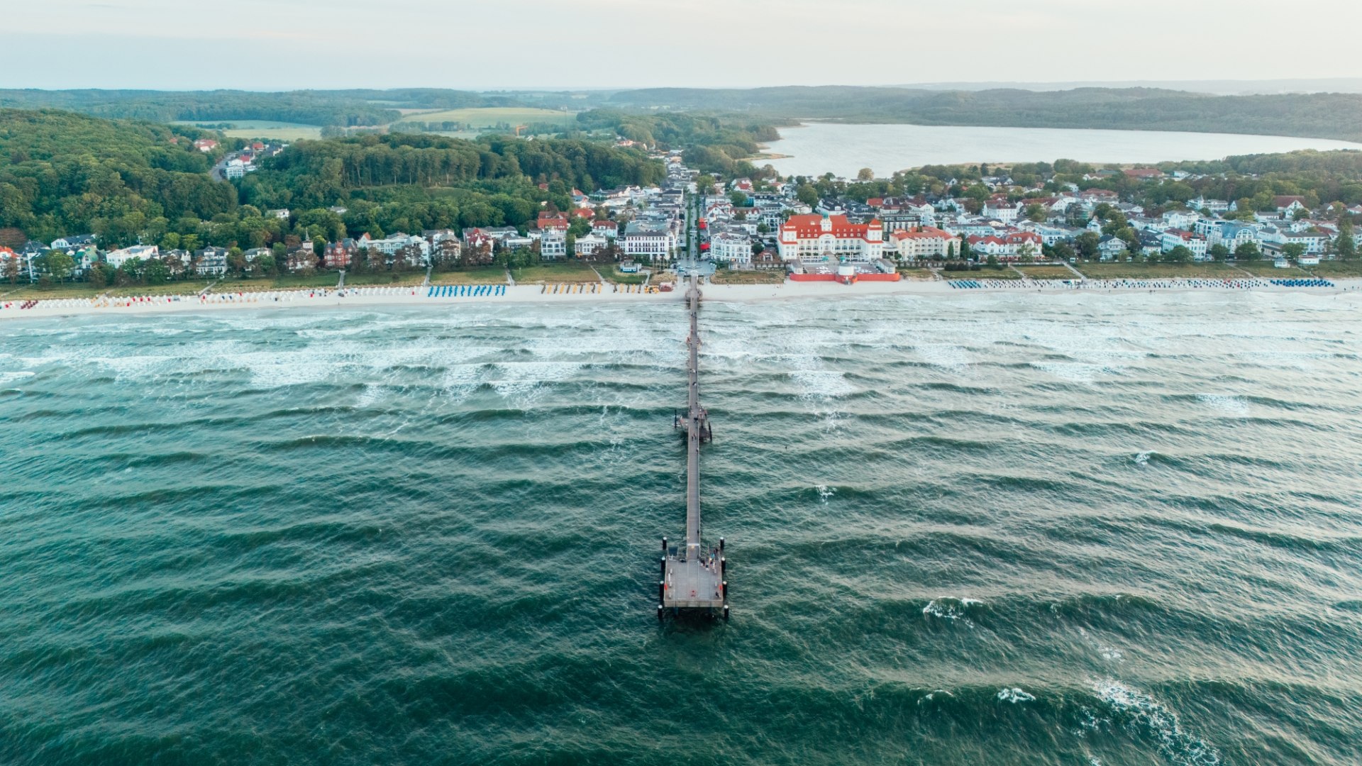 Aerial view of the pier in Binz on the island of Rügen, which juts far out into the Baltic Sea. Behind it lies Binz with its magnificent spa architecture, nestled in a hilly landscape.