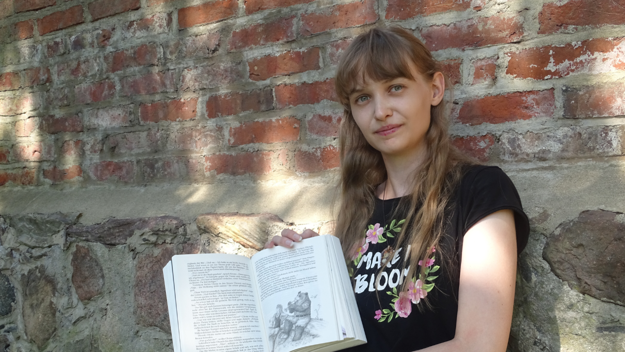 Jose Saefkow sits in a black shirt in front of an old brick wall and shows her book // &copy; Josi Saefkow