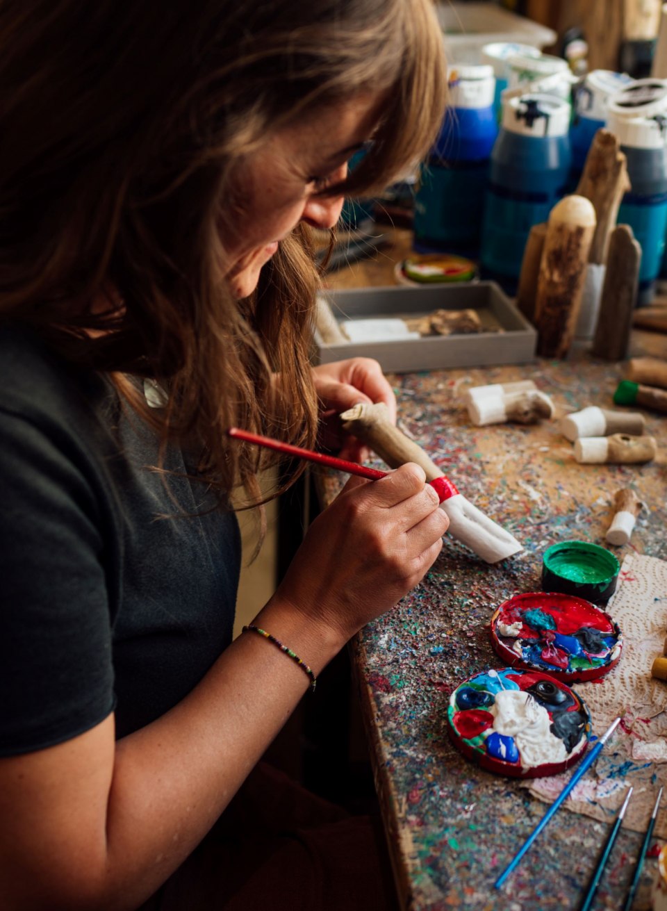 A woman sits at a brightly painted work table and paints a driftwood figure with a brush. On the table are paint pots, brushes and other small driftwood objects that are being artistically decorated. In the background are other handmade figures.