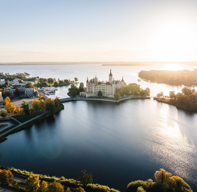 Zamek Schwerin na wyspie na jeziorze o wschodzie słońca, otoczony drzewami i wodą. // Rezydencja na wyspie zamkowej znajduje się pomiędzy wodą a niebem. Poranne światło dotyka wież zamku Schwerin, a jego sylwetka odbija się w spokojnym jeziorze. Historia spotyka się z ogromem - miejsce, które daje przestrzeń do oddychania, a jednocześnie przyciąga wzrok. // © MV-T/Gross Zamek Schwerin na wyspie na jeziorze o wschodzie słońca, otoczony drzewami i wodą.