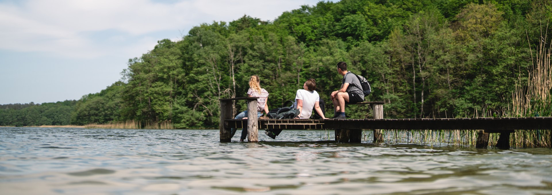 Fancy a break? It's best to stop at every corner and enjoy nature, just like these three friends here on the reedy shore of Lake Groß Labenz near Friedrichswalde., © TMV/Gross 3 friends sit for a break on a jetty on Lake Groß Labenz near Friedrichswalde.