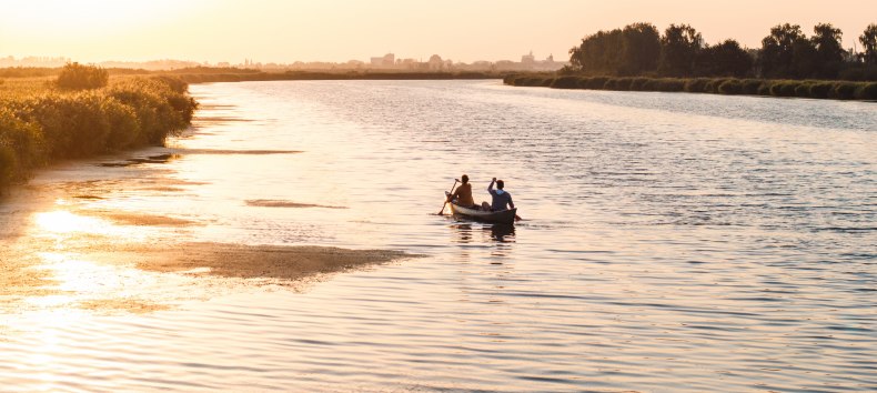 Twee mensen peddelen in een kano over een kalme rivier bij zonsondergang, met gouden licht op het water.