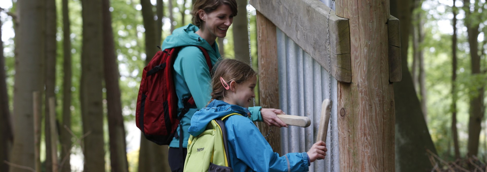 A mother and her daughter stand in front of an installation with sound tubes in the sound forest in Graal-M&uuml;ritz. Both are holding wooden mallets and striking the tubes to produce sounds while smiling.