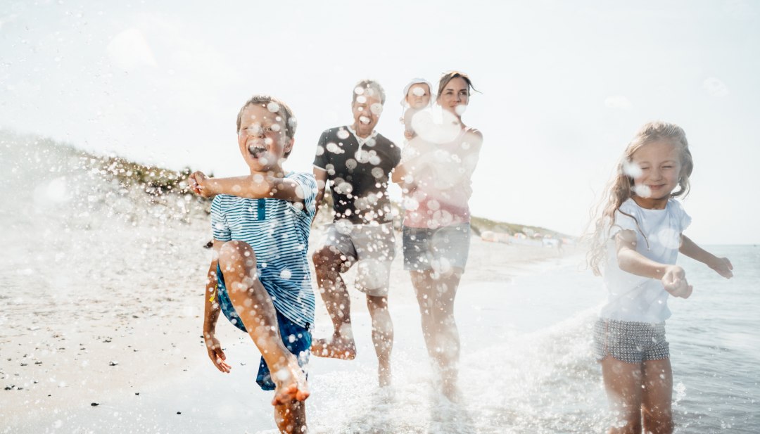 A family runs through the shallow Baltic Sea waves on the beach at Dierhagen, laughing as water splashes vividly in the sun.