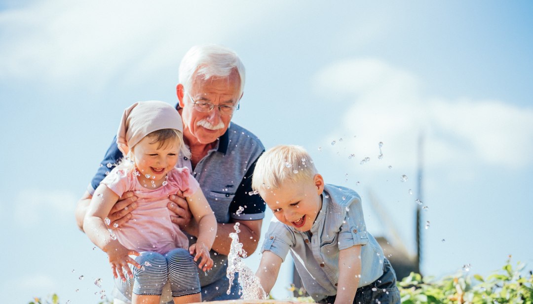 Ein Großvater mit Brille hält ein kleines Mädchen fest, während ein Junge lachend nach einem Wasserstrahl greift // Spritzer, Lachen, Sonnenschein: Im Freilichtmuseum Klockenhagen lassen Kinder mit Opa das Wasser fliegen. // © MV-T/Gaensicke Ein Großvater mit Brille hält ein kleines Mädchen fest, während ein Junge lachend nach einem Wasserstrahl greift // Spritzer, Lachen, Sonnenschein: Im Freilichtmuseum Klockenhagen lassen Kinder mit Opa das Wasser fliegen. // © MV-T/Gaensicke