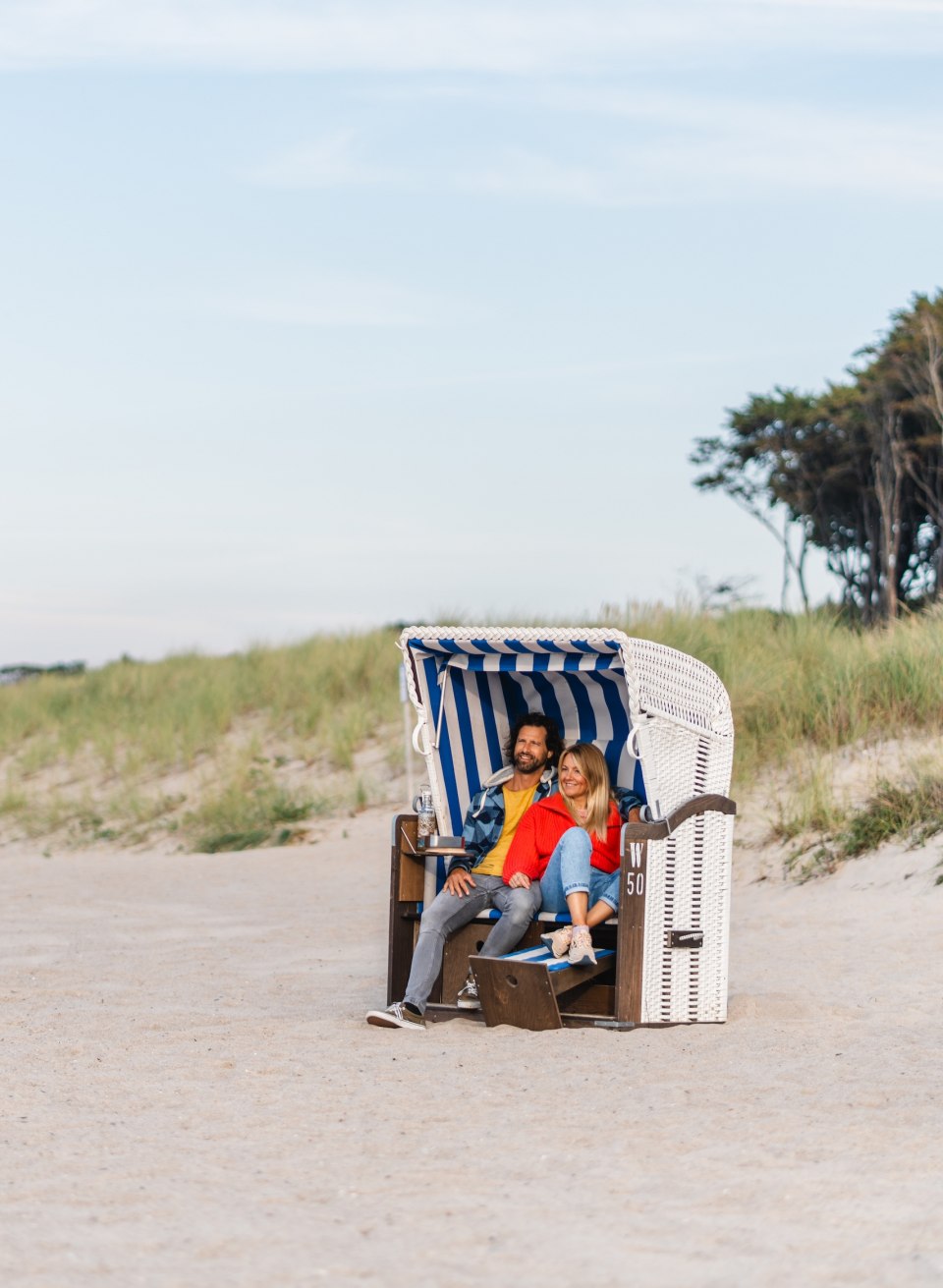 Een koppel zit in een blauw-wit gestreepte strandstoel op het strand in Graal-Müritz, omgeven door zand en duinen met uitzicht op het rustige kustlandschap.