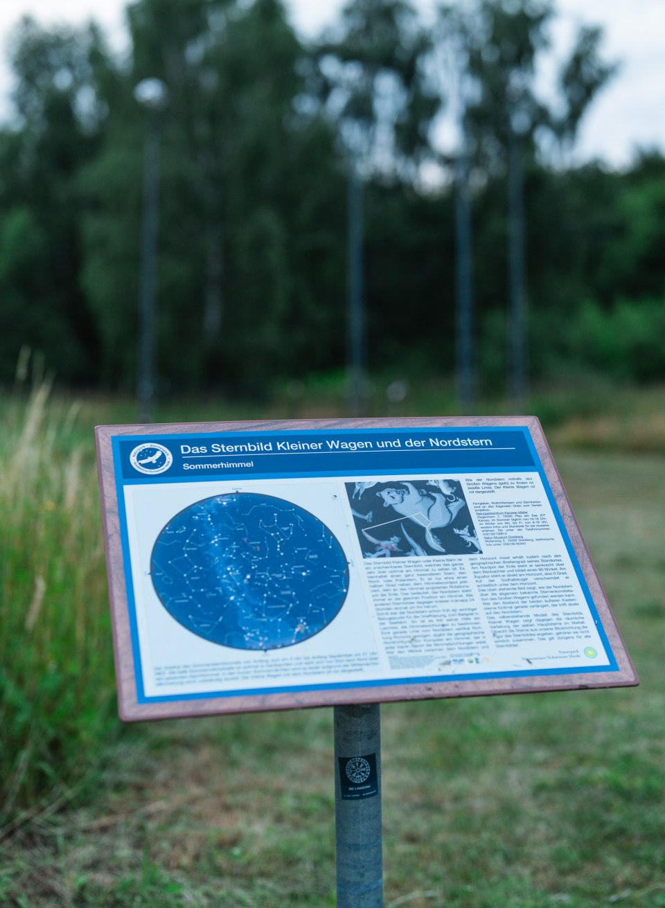 Tablica informacyjna z mapą gwiazd przedstawiającą Wielki Wóz i Gwiazdę Polarną stoi na skraju łąki, a w tle niewyraźnie widać las. // Na tablicy informacyjnej widnieją konstelacje Małego Wozu i Gwiazdy Polskiej – to punkt wyjścia do spędzenia nocy w jednym z ogólnodostępnych miejsc do obserwacji gwiazd w Meklemburgii-Pomorzu Przednim. // © MV-T/Gross Tablica informacyjna z mapą gwiazd przedstawiającą Wielki Wóz i Gwiazdę Polarną stoi na skraju łąki, a w tle niewyraźnie widać las.