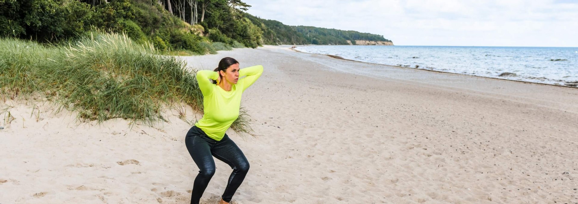 Kobieta na plaży wykonuje ćwiczenia fitness: Przysiady z rękami skrzyżowanymi na karku.