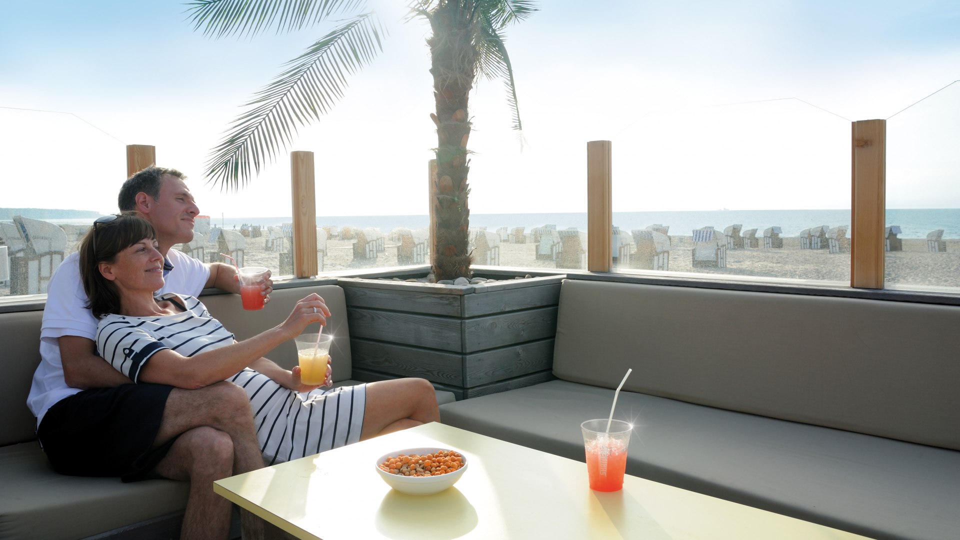 Couple relaxing with drinks on a lounge in the beach bar DÜNE 13 in Warnemünde, with a view of the beach and the Baltic Sea.
