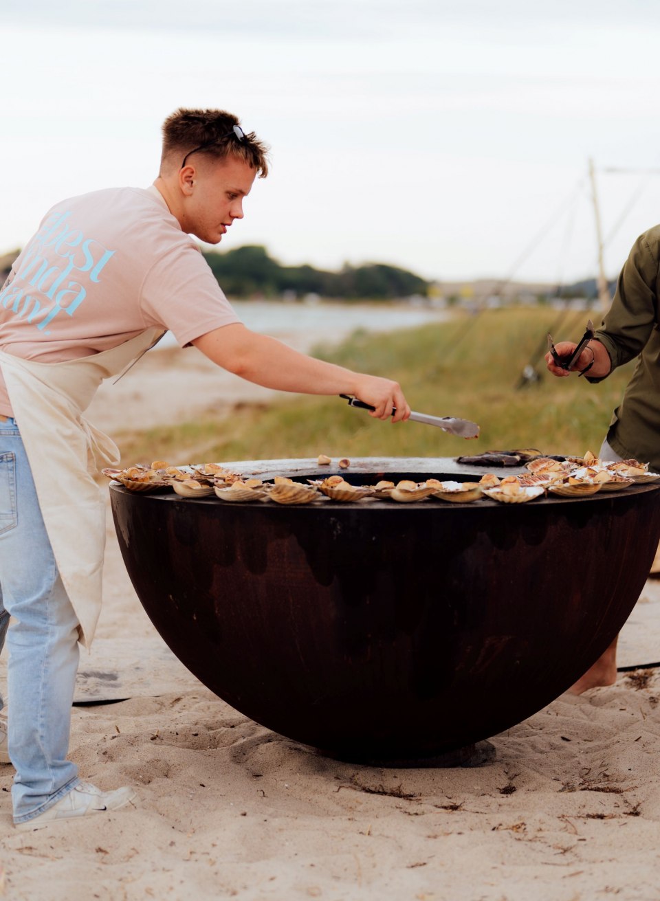 Dwóch mężczyzn przygotowuje regionalne potrawy na grillu na dużej płycie grillowej na plaży w Thiessow; w tle widać morze i namiot. // Na plaży w Thiessow na płytce do smażenia skwierczą świeże, regionalne potrawy – praca zespołowa, dym i słone powietrze. // © MV-T/Petermann Dwóch mężczyzn przygotowuje regionalne potrawy na grillu na dużej płycie grillowej na plaży w Thiessow; w tle widać morze i namiot.