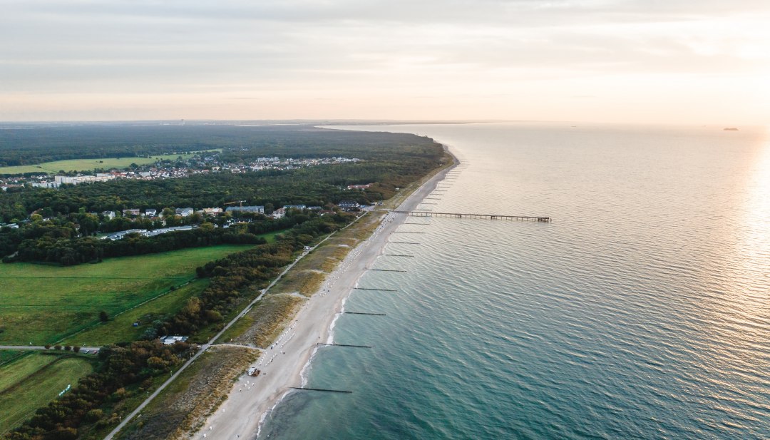 Luchtfoto van de pier en het strand van Graal-Müritz, omgeven door bossen en weilanden, met de Baltische Zee in het licht van een sfeervolle zonsondergang.