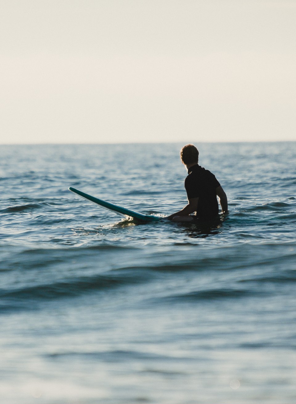A person sits with a surfboard in the gentle Baltic Sea waves off the coast of Warnem&uuml;nde and looks out over the calm water.