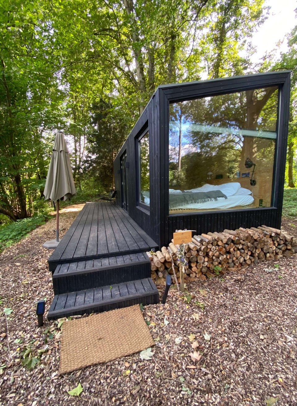 Zwart huisje met een glazen voorgevel midden in een bos bij kasteel Neuhoff, met een stapel hout en terras ervoor.