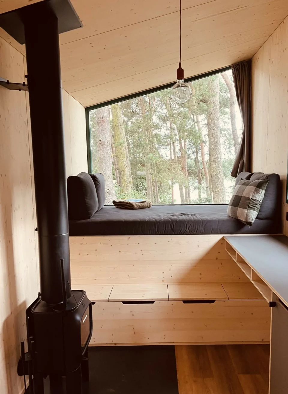 Gezellig interieur van een klein huisje met houten lambrisering, houtkachel en een groot panoramisch raam met uitzicht op het bos.