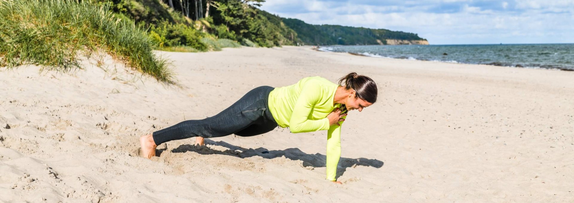 Een vrouw op het strand doet fitnessoefeningen: hier plank met schoudertikken // Steun op je handpalmen. Zodra je een stabiele houding hebt, til je je linkerhand van de grond en breng je hem naar je rechterschouder. Tik één keer en dan terug op het strandzand. Zorg ervoor dat je heupen en lichaam niet opzij bewegen wanneer je op je schouders tikt. Alleen de hand moet naar de tegenoverliggende schouder bewegen. 6-10 herhalingen per kant // © MV-T/Tiemann Een vrouw op het strand doet fitnessoefeningen: hier plank met schoudertikken