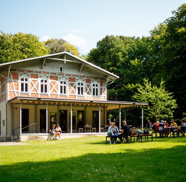 A bright building with a terrace, in front of which several people are sitting on benches on a green lawn. // &copy; Nationalpark-Zentrum K&Ouml;NIGSSTUHL / Dan Petermann