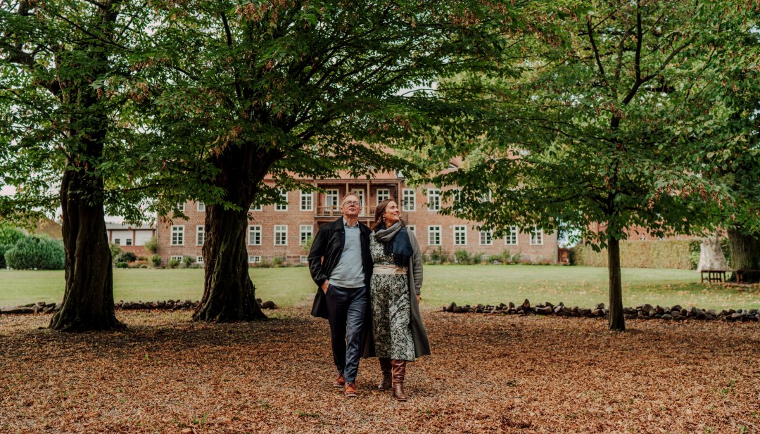 Een koppel wandelt onder grote bomen in het park, met het Romantik Hotel Gutshaus Ludorf op de achtergrond. // Haal diep adem onder oude bomen, verzamel je stappen, met het landhuis van Ludorf in zicht - rust met de uitgestrektheid van Mecklenburg. // © MV-T/Petermann Een koppel wandelt onder grote bomen in het park, met het Romantik Hotel Gutshaus Ludorf op de achtergrond.