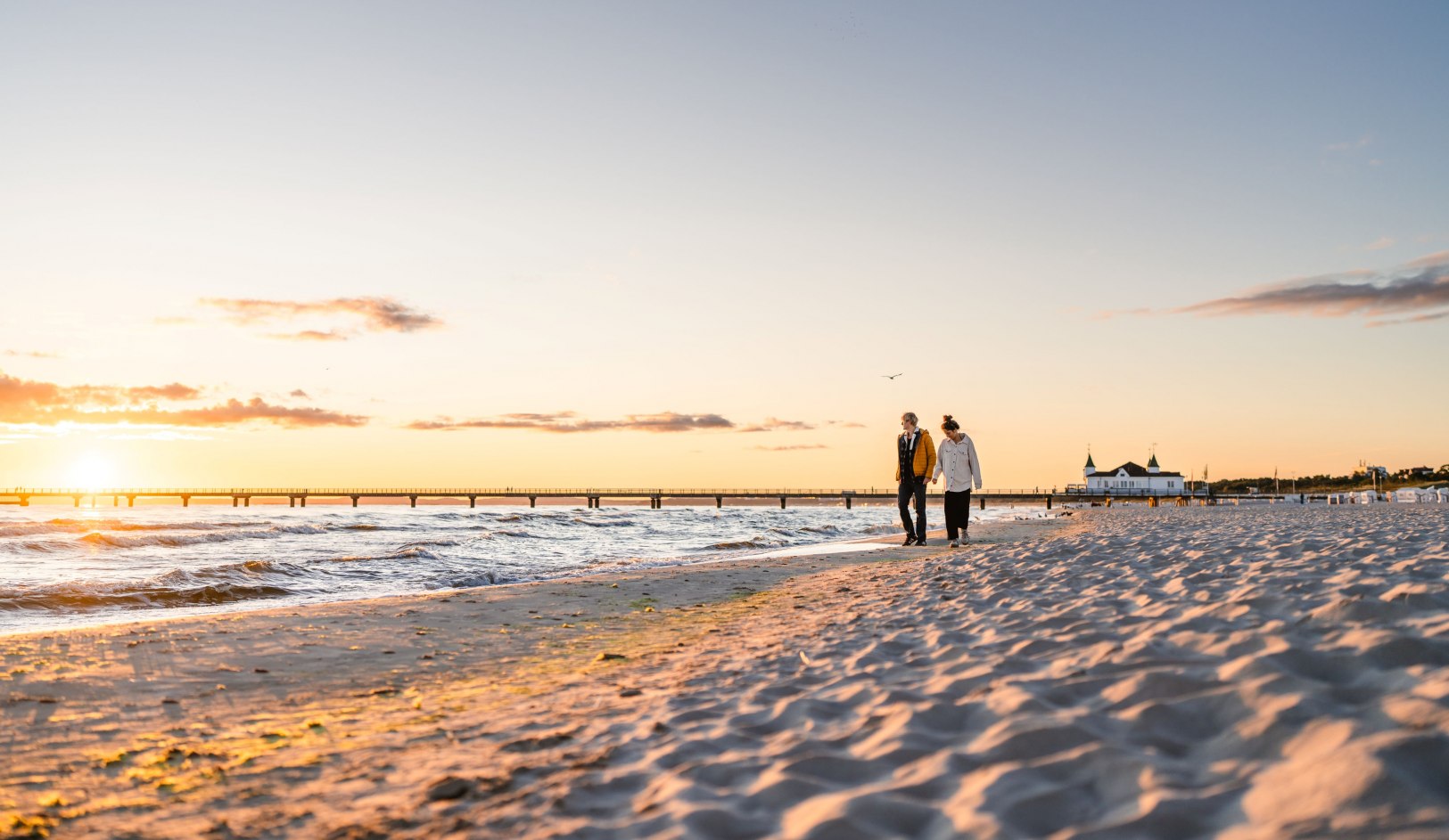 Paar aan het wandelen op het strand in Ahlbeck bij zonsondergang met de pier op de achtergrond. // Gouden licht baadt het strand van Ahlbeck in warme kleuren. De pier steekt uit in zee terwijl zachte golven het fijne zand beroeren. Een moment tussen dag en avond in het keizerlijke resort van Usedom dat uitnodigt om even stil te staan. // © MV-T/Gross Paar aan het wandelen op het strand in Ahlbeck bij zonsondergang met de pier op de achtergrond.