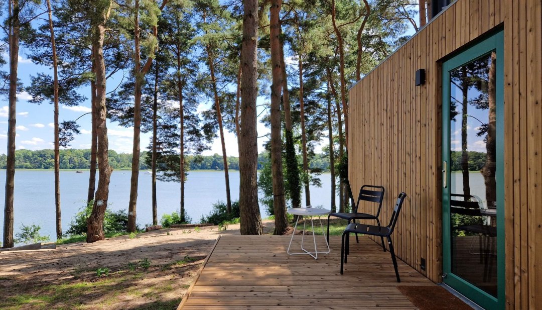 Houten terras van een klein huisje met uitzicht op het meer, omringd door pijnbomen in een rustige natuurlijke omgeving.