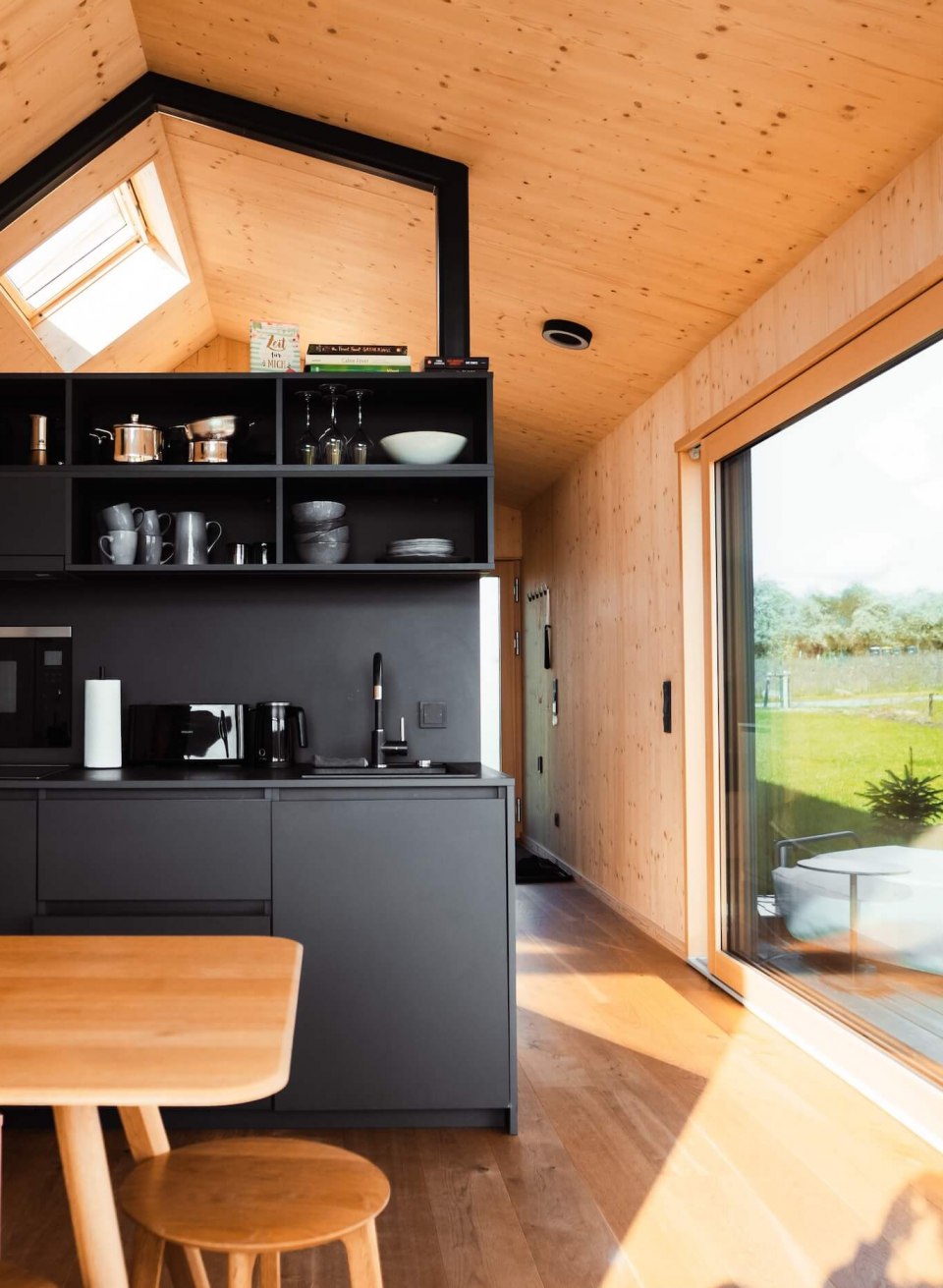 Modern Tiny House met zwarte kitchenette, houten meubels en een grote schuifdeur naar het terras.