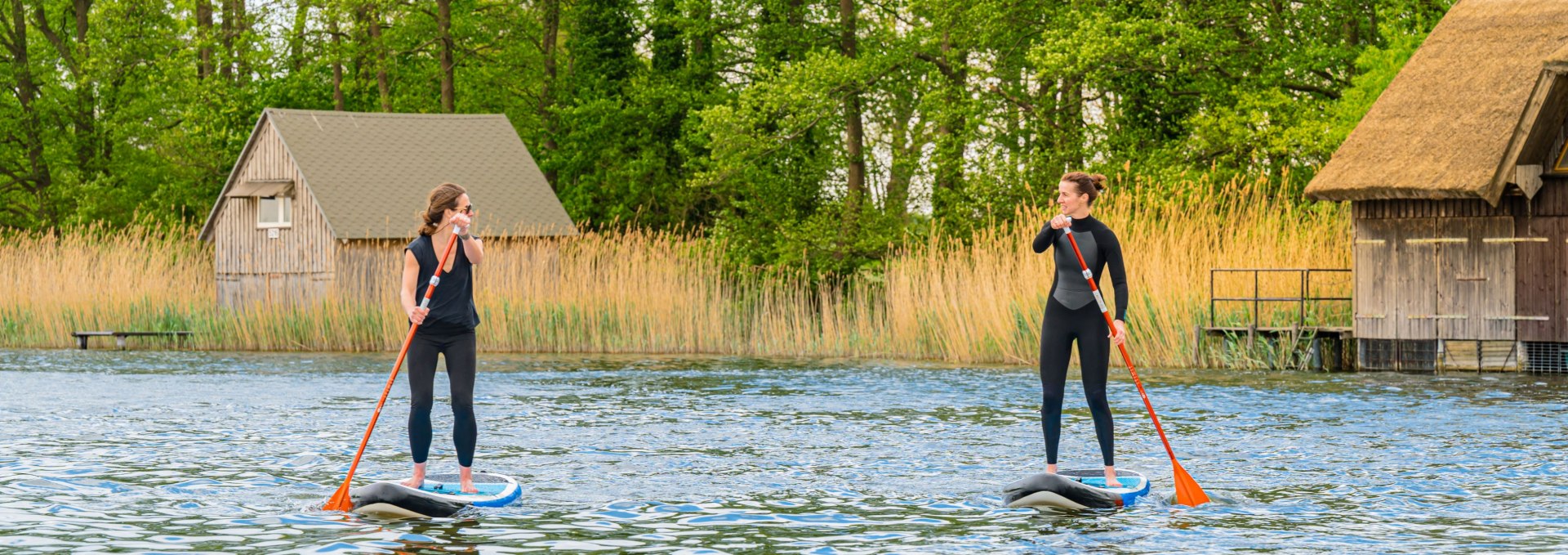 To kvinder, der st&aring;r p&aring; paddleboards foran str&aring;t&aelig;kkede b&aring;dehuse og t&aelig;tte r&oslash;rskove i Mecklenburgs s&oslash;omr&aring;de.