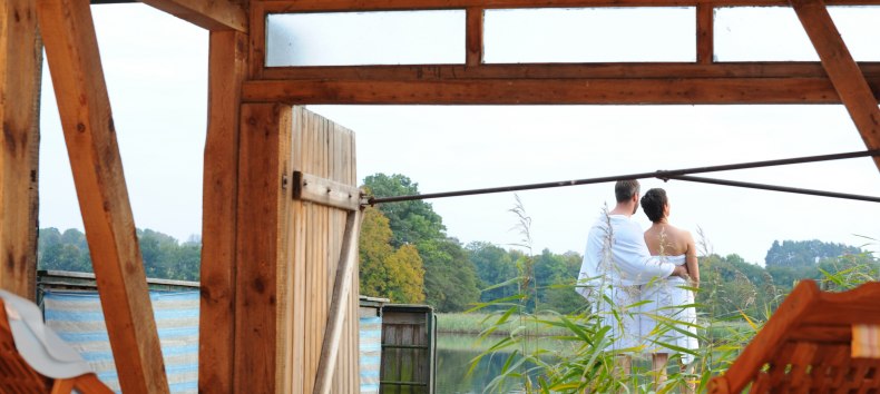 A couple in towels stands on a terrace by the lake, surrounded by reeds, with a view of the water in the Feldberg lake landscape. Wooden loungers can be seen in the foreground.
