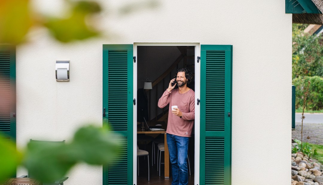 Man aan de telefoon met een koffiekopje in zijn hand voor de deur van een vakantiehuisje op het eiland Poel tijdens zijn werkvakantie.