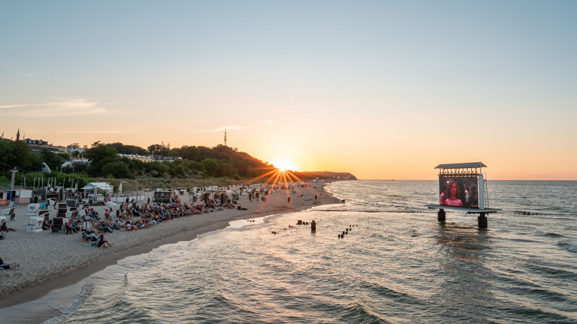 Kino plażowe na wyspie Uznam z ekranem LED w morzu i zachodem słońca za widzami na plaży nad Morzem Bałtyckim.