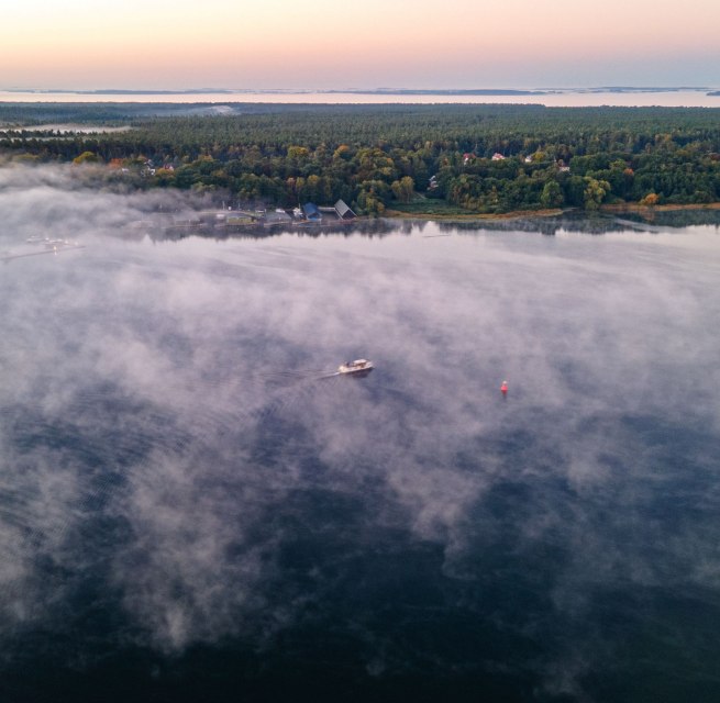 Luchtfoto van de Müritz in de vroege ochtend met mistflarden waar een boot langzaam doorheen vaart.