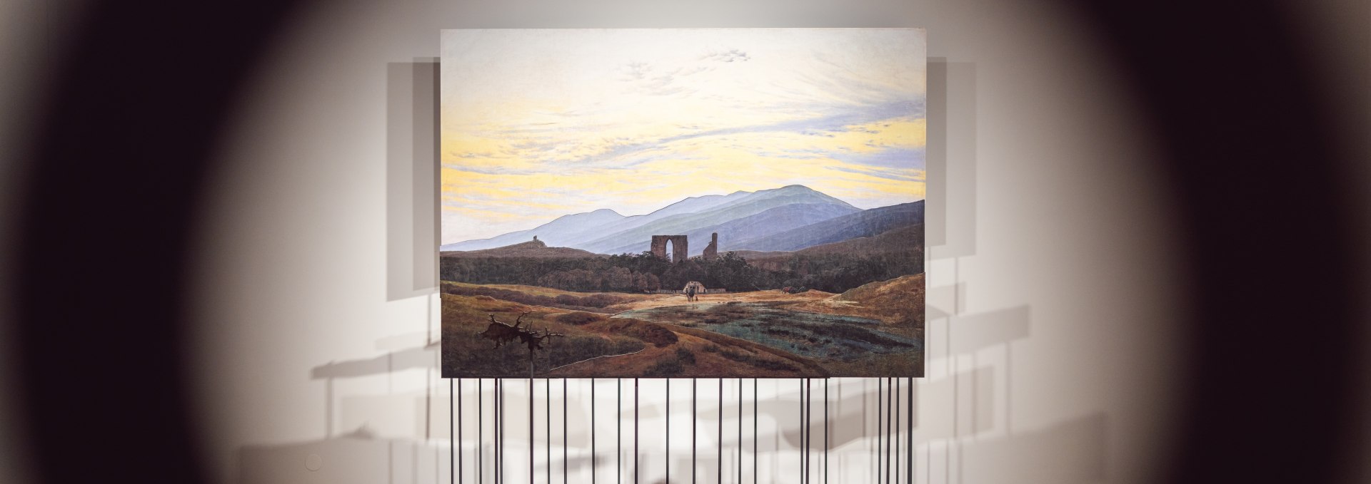 Widok obrazu pejzażowego Caspara Davida Friedricha w Pomorskim Muzeum Krajowym w Greifswaldzie otwiera się przez dziurkę od klucza. Zainscenizowana perspektywa wzmacnia intymność spotkania z dziełem sztuki i zachęca do bliższego przyjrzenia się. Moment pomiędzy dystansem a bliskością., © MV-T/Gross Romantyczny obraz pejzażowy na cokole, sfotografowany przez okrągły otwór, w Państwowym Muzeum Pomorskim.